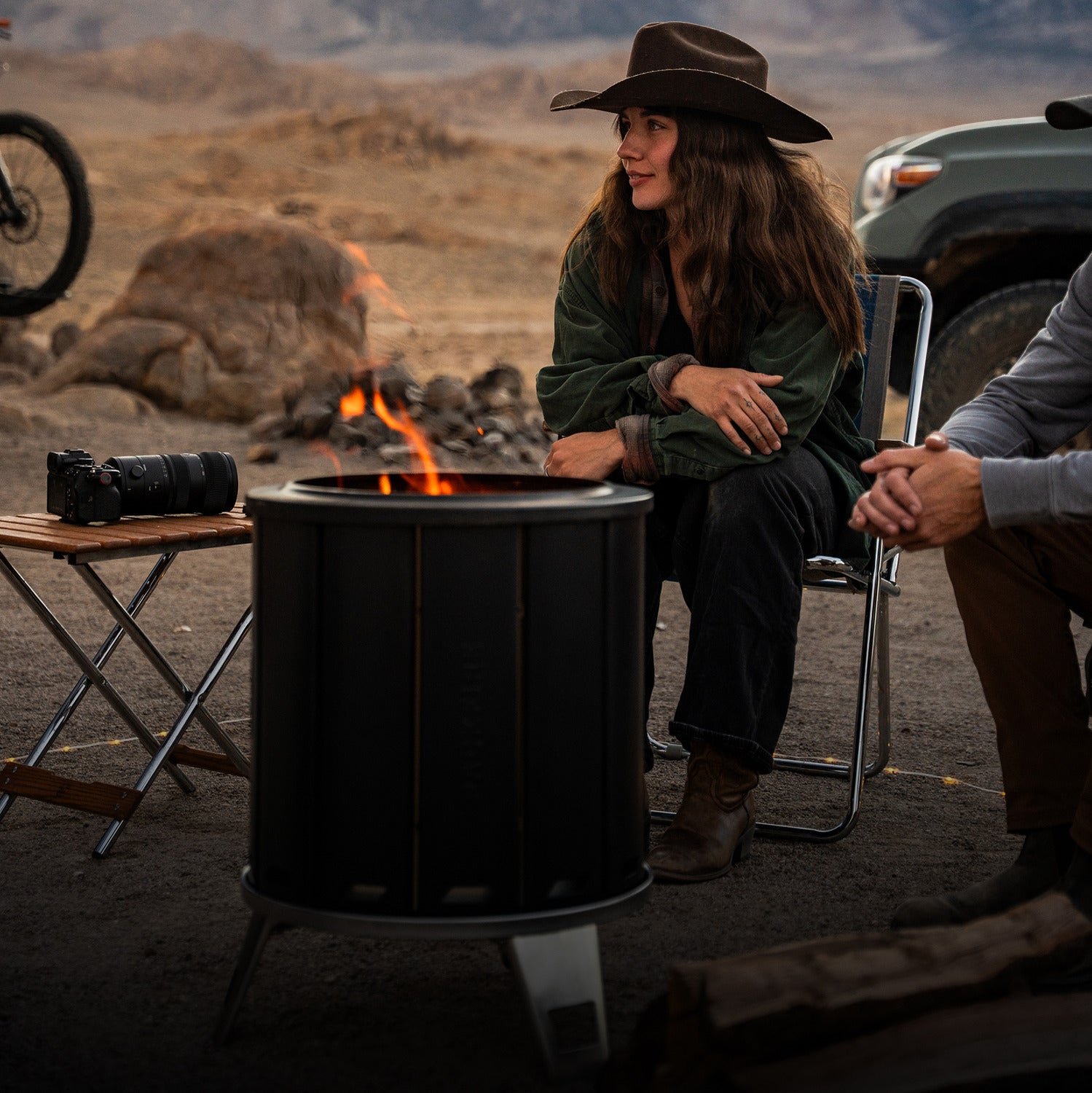 Two people sitting by a fire pit in a desert setting with a motorcycle and vehicle in the background.