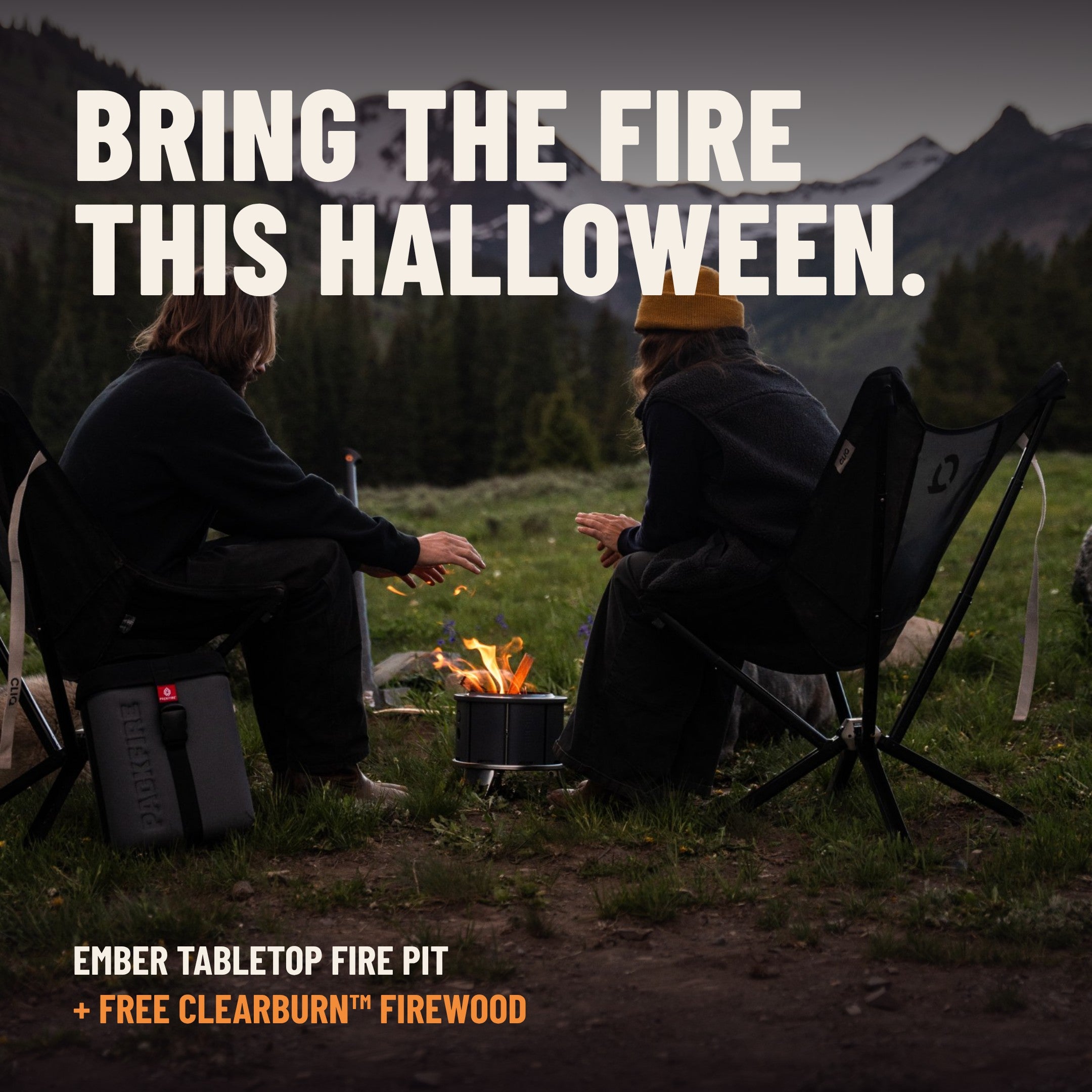 Two friends warming their hands over the Ember fire pit at dusk below snowy mountains, with the text Bring the Fire This Halloween, Ember tabletop fire pit plus free ClearBurn Tabletop Firewood