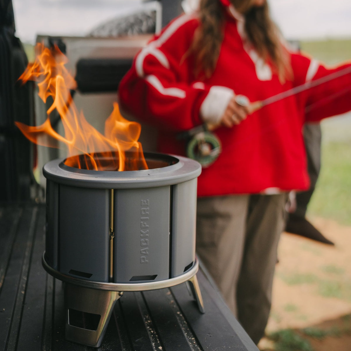Ember fire pit burning on a truck tailgate while a woman in a red jacket holds a fly rod in the background