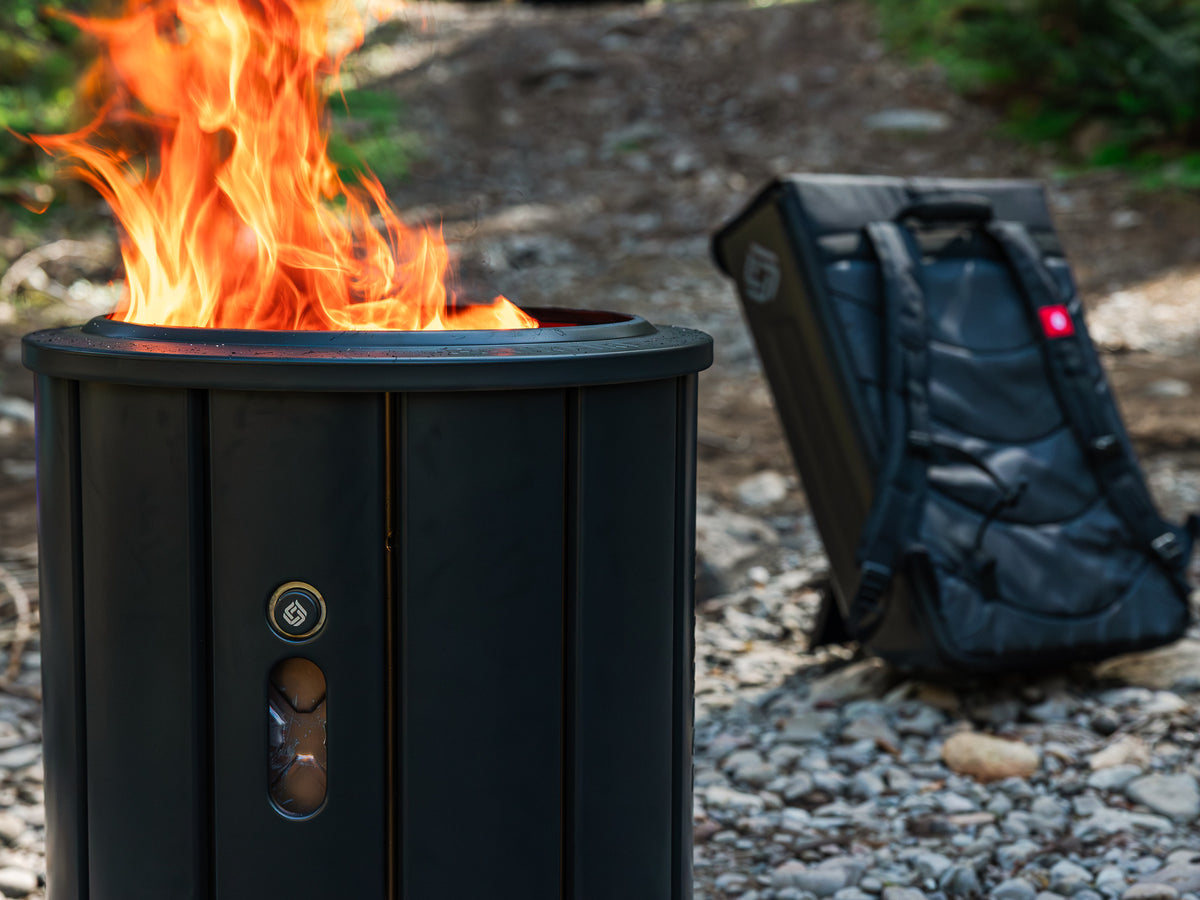 Black fire pit with flames and a black backpack in the background on a gravel surface.